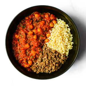 A black bowl with Engine 38 Tortellini, chickpea tomato stew, yellow rice, and ground meat in three separate sections.