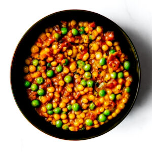 A black bowl of Engine 38 Tortellini, a tomato-based stew with yellow lentils and green peas, seen from above on a white background.