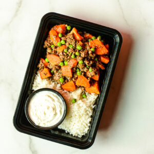 The Flexican Fiesta features ground beef, carrots, peas, white rice, and white sauce in a black tray, set on a marble surface.