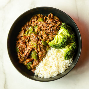A black bowl of Chicken & Rice 9000 features shredded chicken, green peppers, steamed white rice, and broccoli florets arranged in separate sections.