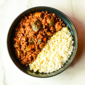 A bowl of Chicken & Rice 9000 with white rice and hearty red chili sits on a light surface.