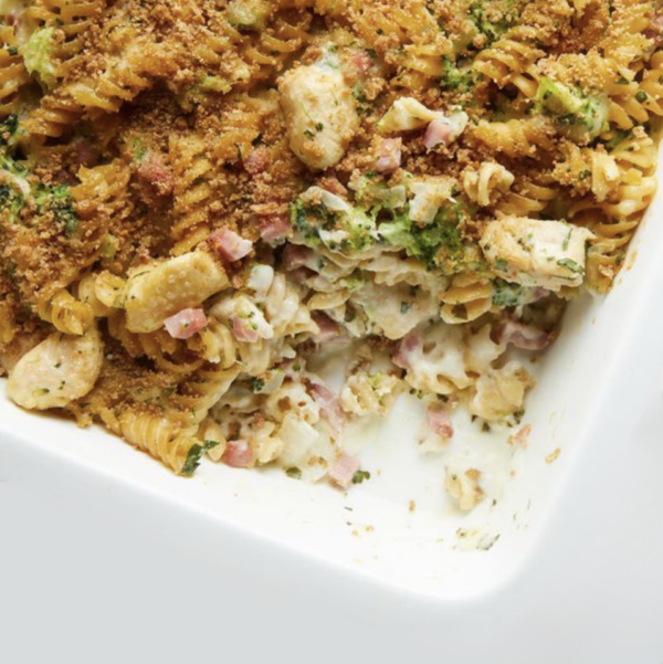 A close-up view of Chicken Cordon Bleu Tetrazzini in a white dish, featuring fusilli pasta, chicken pieces, ham, broccoli, and a crispy breadcrumb topping. A portion in the corner of the dish is missing, revealing the cheesy interior.