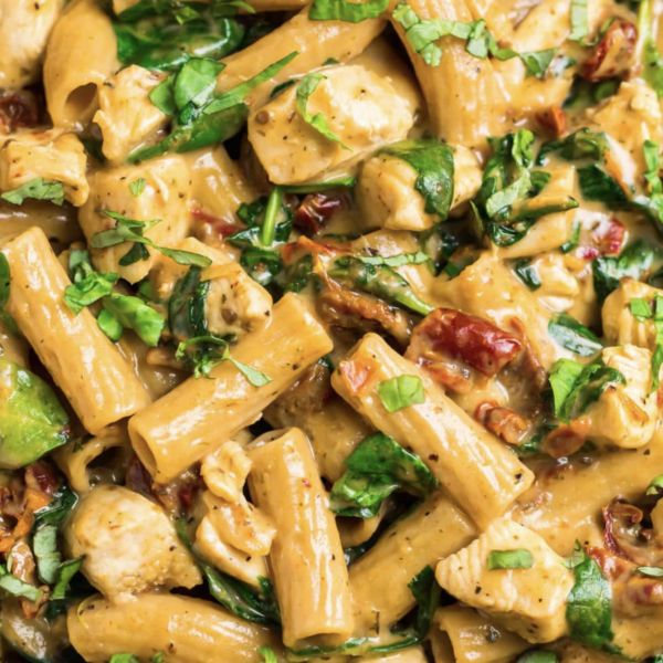 A close-up of Tuscan Mac & Cheese featuring creamy pasta with rigatoni, tender pieces of chicken, sun-dried tomatoes, and spinach. The dish is garnished with fresh basil, creating a colorful and appetizing presentation.