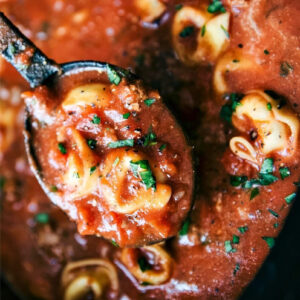 Close-up of a ladle lifting cheese tortellini topped with The GOD Mother tomato sauce, garnished with chopped parsley. The sauce is thick and speckled with visible herbs and spices.