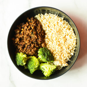 A black bowl of Pardon My French… Clucker with ground meat, steamed broccoli florets, and cooked brown rice sits on a white surface.