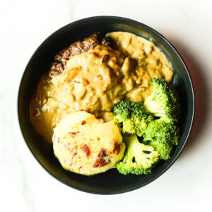 The Pardon My French… Clucker features a black bowl with mashed potatoes, broccoli florets, and a hamburger patty topped with brown gravy.