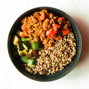 A bowl of Pardon My French… Clucker with cooked quinoa, sautéed zucchini, and ground meat with red bell peppers is arranged in sections on a white background.