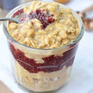 A glass jar packed with delectable PBJ Oats, featuring layers of creamy oats and chia seeds. A spoon is dipped into the top layer, showcasing a striking blend of oats and red chia seeds that form a textured and vibrant design, capturing the essence of a classic peanut butter and jelly snack.