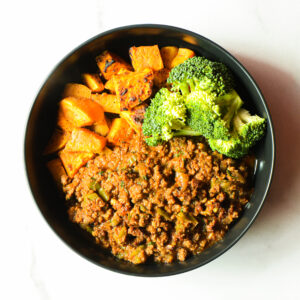 A black bowl of Pardon My French… Clucker with ground meat in sauce, steamed broccoli florets, and roasted sweet potato cubes on a white surface.
