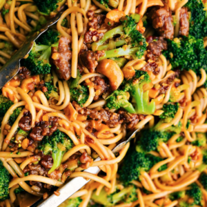 A close-up of a vibrant, colorful stir-fry featuring noodles, broccoli florets, and pieces of chicken, all tossed in a rich, glazed sauce with sliced almonds scattered throughout. Tongs are visible, ready to serve the dish.