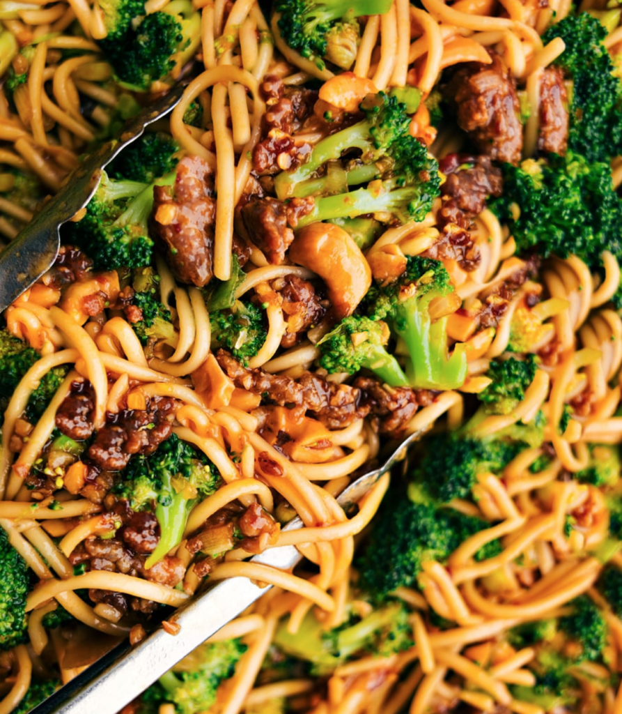A close-up of a vibrant, colorful stir-fry featuring noodles, broccoli florets, and pieces of chicken, all tossed in a rich, glazed sauce with sliced almonds scattered throughout. Tongs are visible, ready to serve the dish.