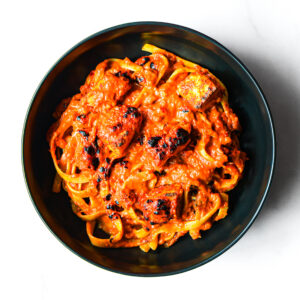 Alfredo del Bombero: Grilled protein over pasta with creamy red sauce, served in a black bowl on a white surface.