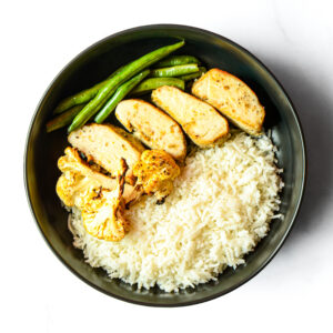 A bowl of Alfredo del Bombero with white rice, sliced chicken breast, green beans, and roasted cauliflower on a white background.