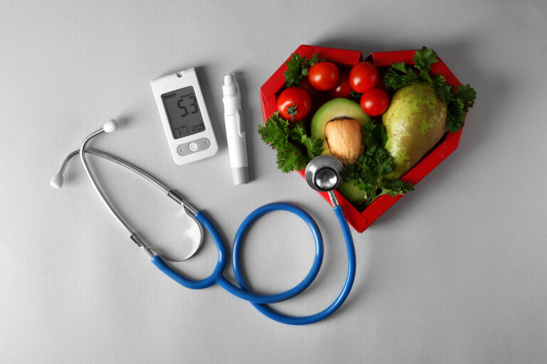 A stethoscope, glucometer, and lancing device sit by a heart-shaped dish of vegetables—highlighting senior meal delivery for chronic conditions on a gray background.