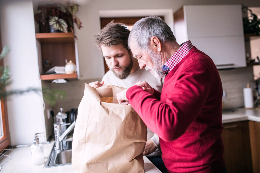 An older man and a younger man look into a large brown paper bag together in a kitchen, reviewing their senior meal delivery checklist.