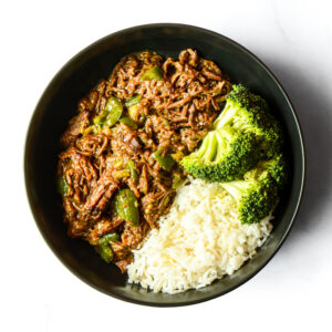 The Nacho Libre Gains Bowl features shredded beef with green peppers in sauce, served with white rice and steamed broccoli.