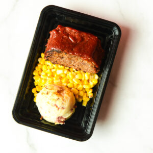 A black tray containing a slice of meatloaf with ketchup glaze, a serving of corn, and a scoop of mashed potatoes on a white surface.
