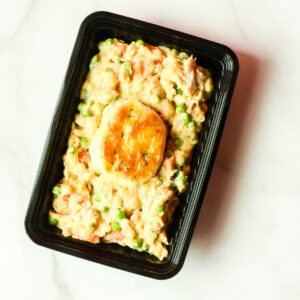 A black plastic tray contains a serving of chicken pot pie filling with peas and carrots, topped with a single round biscuit.