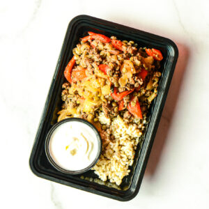 The Angry Korean meal features cooked ground beef, vegetables, brown rice, and white sauce in a black tray on a white surface.