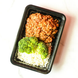 The Angry Korean: a black plastic container with white rice, steamed broccoli, and seasoned ground beef in tomato sauce on a white surface.