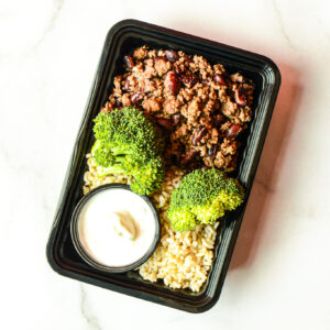 The Angry Korean: A meal container with ground meat, beans, steamed broccoli, brown rice, and a small cup of white sauce on a white surface.