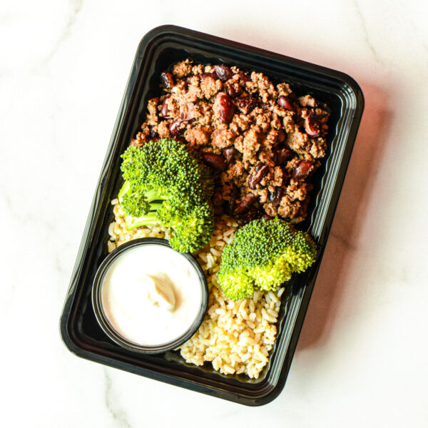 The Angry Korean: A meal container with ground meat, beans, steamed broccoli, brown rice, and a small cup of white sauce on a white surface.