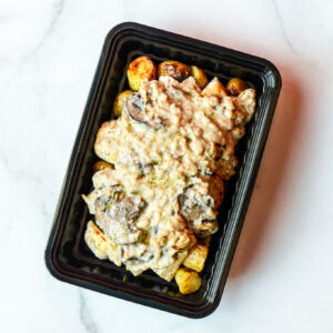 A black plastic container of The Angry Korean's roasted potatoes topped with creamy mushroom sauce on a white surface.
