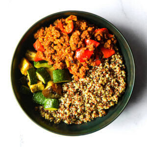 A bowl of Jack'd-Atouille featuring ground meat, red bell peppers, roasted zucchini, and quinoa sits on a white surface.
