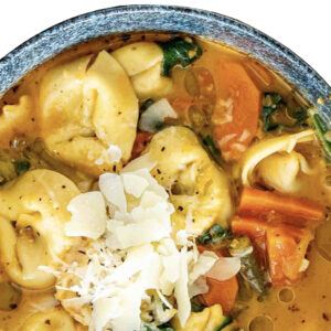A close-up of Pasta La Vista, Veggies tortellini soup topped with carrots, spinach, and shaved parmesan cheese.