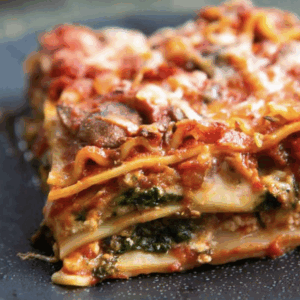 A close-up of Veg Me Hard Lasagna, revealing layers of pasta, spinach, tomato sauce, mushrooms, and melted cheese on a dark plate.