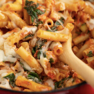 Close-up of No Balls Ziti pasta with melted cheese, tomato sauce, and spinach, being stirred with a wooden spoon.