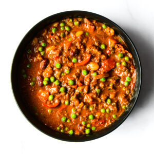 Korma Kommander is a thick stew with ground meat, green peas, kidney beans, and tomato chunks in a reddish-brown sauce.