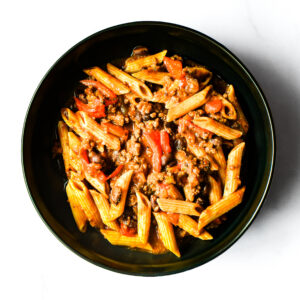 A black bowl filled with Korma Kommander penne pasta, ground meat, tomato sauce, diced tomatoes, and black beans on a white surface.