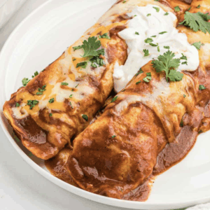 Two Holy Hell! Wheres The Cow? enchiladas topped with cheese and sauce, garnished with cilantro and sour cream on a white plate.