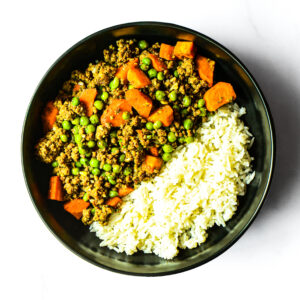 A bowl of Korma Kommander with cooked white rice, ground meat, green peas, and diced carrots.