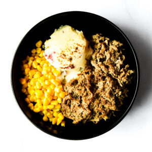 The Korma Kommander shows a black bowl with corn, mashed potatoes, and shredded meat against a white background.