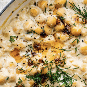 A close-up of Savage Chickpeas, creamy and garnished with fresh dill, black pepper, and a drizzle of oil in a white dish.
