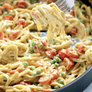 A fork lifts Veg Me Hard, Alfredo creamy pasta with cherry tomatoes and green peas from a skillet.
