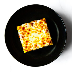 A square slice of The F*d Up Fajita with browned cheese on top, served on a black plate against a white background.