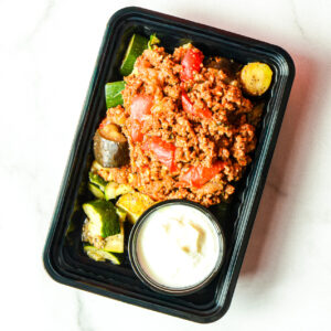 A black meal tray with cooked ground meat and tomatoes over chopped vegetables, accompanied by a small container of white sauce.