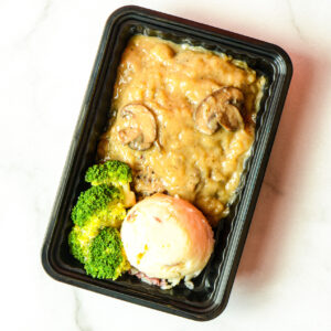 A black plastic tray with mashed potatoes, steamed broccoli, and a meatloaf slice topped with brown gravy and mushrooms.