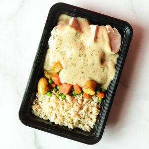 A meal tray containing sliced chicken with creamy sauce, roasted potatoes, mixed vegetables, and a serving of brown rice.