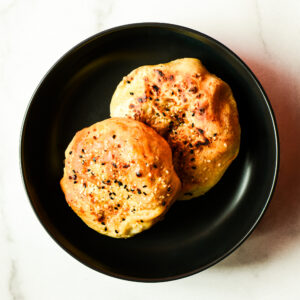 Two golden-brown, seeded buns from "Cheesus! Is That Ham In Your Buns? - WL" are served on a black plate over a white surface.