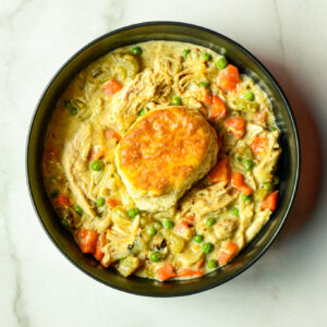 A bowl of creamy chicken pot pie filling with carrots, peas, chicken, and a biscuit on top, served on a white marble surface.