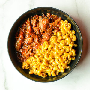 A black bowl containing macaroni and cheese on one side and pulled barbecue meat on the other, placed on a white surface.