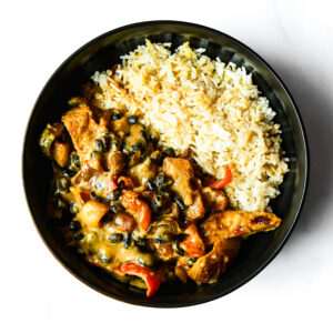 The F*d Up Fajita: A black bowl with white rice and creamy chicken-vegetable stew with capers, viewed from above on a white surface.