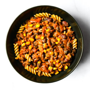 A black bowl holds The F*d Up Fajita, featuring rotini pasta topped with a chunky tomato sauce, beans, corn, and vegetables.