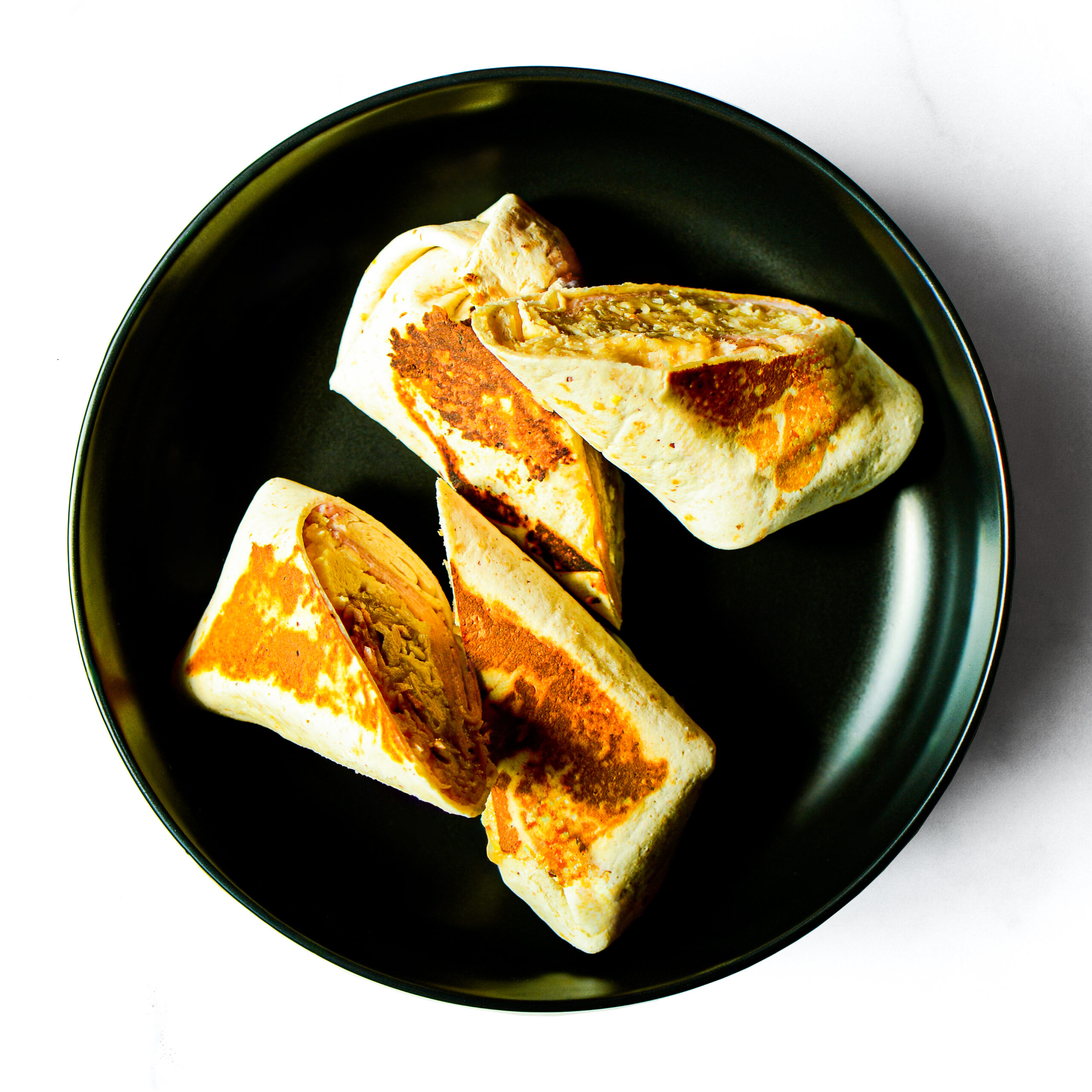 A black plate holds three Pork'in The Morning burritos, grilled with golden brown spots; one is cut in half to reveal the filling.