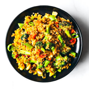 A black plate with The F*d Up Fajita—quinoa, broccoli, green onions, mushrooms, and red bell peppers—arranged on a white surface.