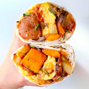 A hand holds a halved breakfast burrito filled with scrambled eggs, potatoes, beans, orange sweet potato slices, and garnished with chopped herbs.